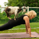 Woman in black yoga clothes smiles as a goat climbs on her back while she planks on a yoga mat outside.