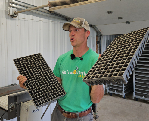 A man holds up empty seedling trays in each hand while speaking.