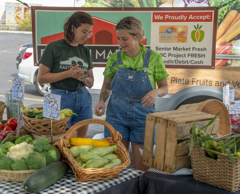 two people stand behind displays of vegetables at a farmers market booth.