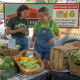 two people stand behind displays of vegetables at a farmers market booth.