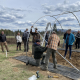 A crowd watches while two people assemble the frame of a high tunnel outdoors